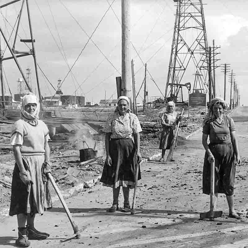 Schwarz-Weiß-Fotografie von Peter Bock-Schroeder aus dem Jahr 1956: Vier Frauen in traditioneller Kleidung mit Kopftüchern posieren auf einer unbefestigten Straße inmitten eines Aserbaidschanischen Ölfeldes, umgeben von stählernen Bohrtürmen und Stromleitungen. Sie halten Werkzeuge wie Spitzhacken und Schaufeln, im Hintergrund ist ein kochender Topf zu sehen.