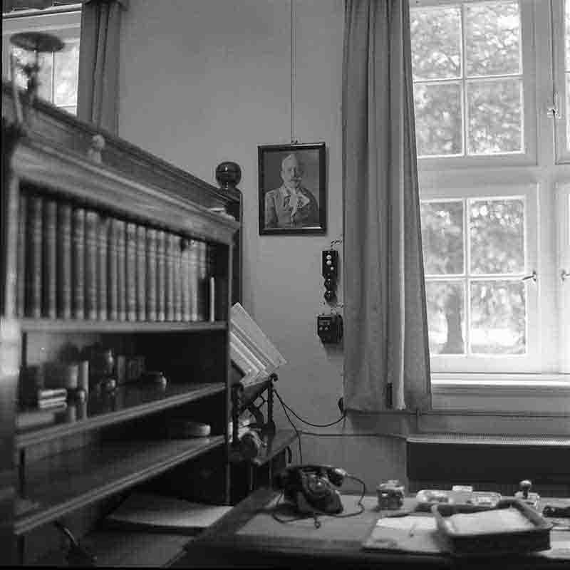 Historische Innenaufnahme von Haus Doorn, fotografiert von Peter Bock-Schroeder (1960). Gezeigt wird ein Arbeitszimmer mit einem dunklen Holzregal voller Bücherreihen und einem Schreibtisch im Vordergrund, auf dem ein altes Wählscheibentelefon steht. An der Wand hängt ein Porträt von Kaiser Wilhelm II. in Uniform.