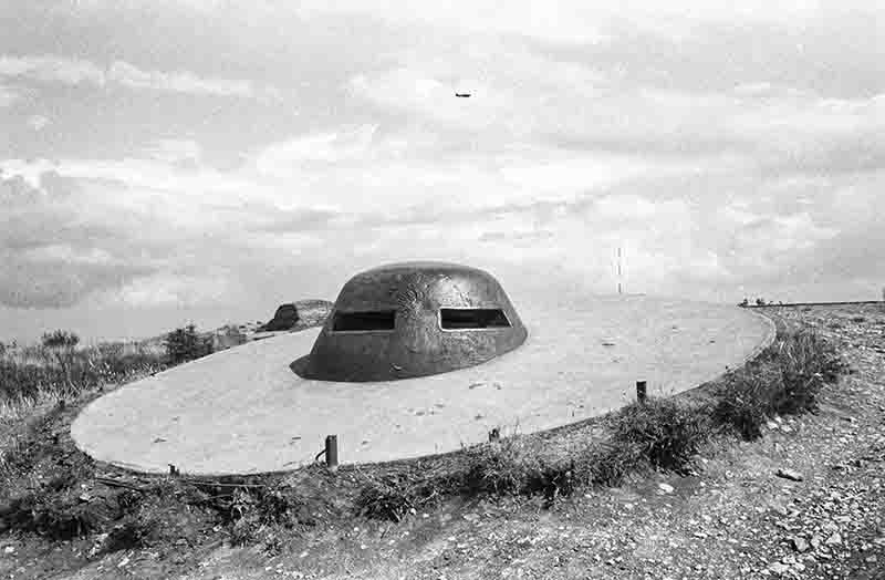Historische Schwarz-Weiß-Aufnahme einer stählernen Panzerkuppel (Beobachtungsglocke) eines deutschen Bunkers am Atlantikwall in der Normandie, eingebettet in einen runden Betonkragen unter bewölktem Himmel mit einem Flugzeug in der Ferne.