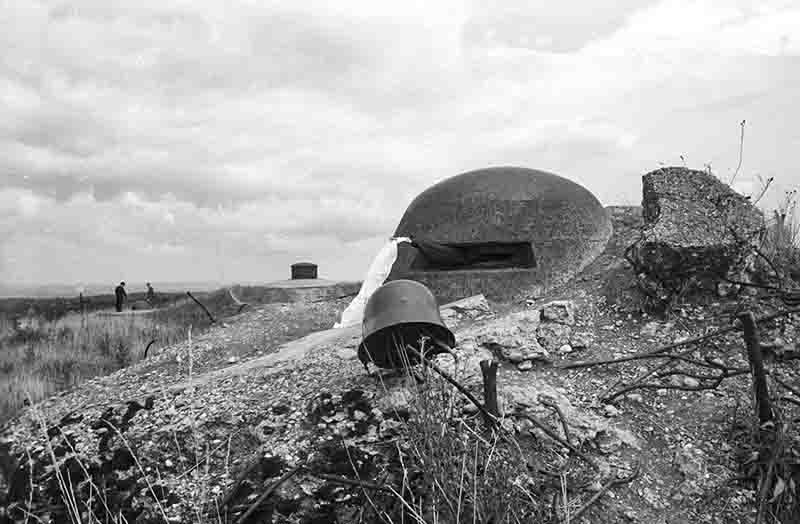 Historische Schwarz-Weiß-Aufnahme an einer Atlantikwall-Stellung in der Normandie. Im Vordergrund liegt ein verlassener deutscher Stahlhelm auf dem Erdboden. Dahinter ist eine betonierte Bunkerkuppel mit Sehschlitz zu sehen, aus dem ein weißes Tuch hängt. Im Hintergrund eine Hügellandschaft mit zwei Personen.