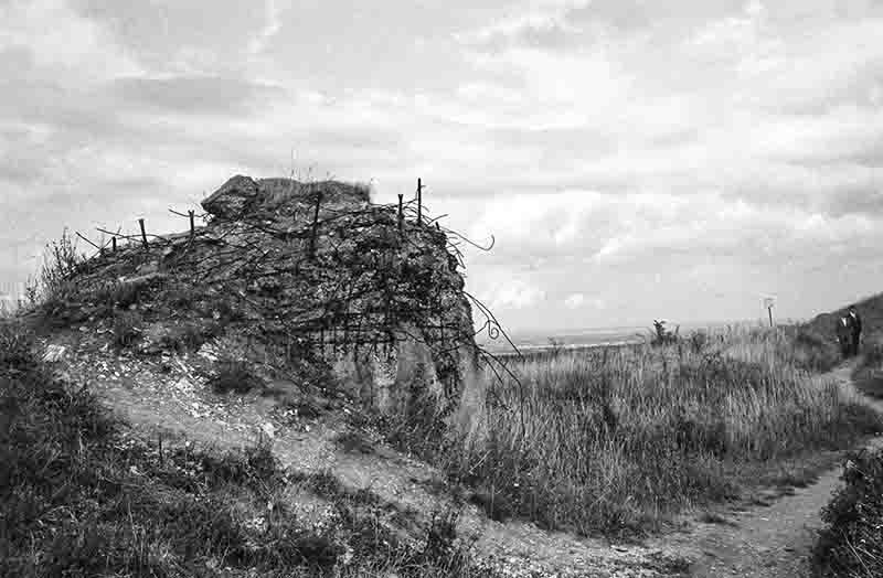 Historische Schwarz-Weiß-Aufnahme eines stark zerstörten Betonbunkers des Atlantikwalls in der Normandie. Der Beton ist zertrümmert, und verbogene Bewehrungsstähle (Armierungseisen) ragen wild aus den Trümmern. Im Hintergrund eine grasbewachsene Klippenlandschaft mit Spaziergängern unter bewölktem Himmel.