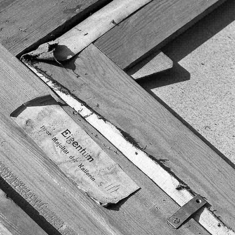 Nahaufnahme einer historischen Transportkiste oder eines Bilderrahmen-Rückens aus Holz im Haus Doorn, fotografiert von Peter Bock-Schroeder (1960). Ein beschädigtes Papieretikett trägt die Aufschrift „Eigentum Ihrer Majestät der Kaiserin“. Die Schwarz-Weiß-Aufnahme betont die raue Textur des Holzes und das abblätternde Papier.