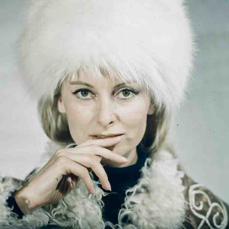 Farbporträt von Eliette von Karajan, die eine große, weiße Fellmütze trägt und den Zeigefinger an die Unterlippe hält. Sie blickt direkt in die Kamera mit einem sanften, nachdenklichen Ausdruck.