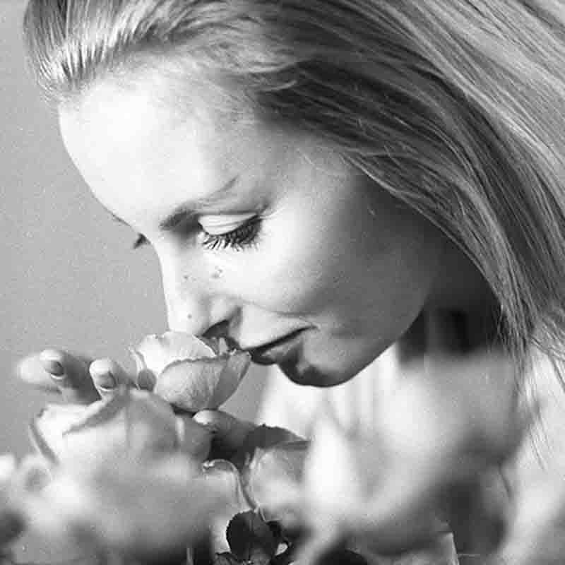 Eine Schwarz-Weiß-Nahaufnahme von Eliette von Karajan, die mit geschlossenen Augen an einer Rose riecht und die Blume sanft an ihre Nase hält. Ihr blondes Haar fällt über ihre Schultern.