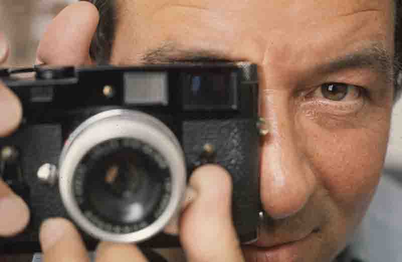 Nahaufnahme vom Fotografen Peter Bock-Schroeder, der eine schwarze Messsucherkamera vor sein Gesicht hält und durch den Sucher direkt in die Kamera blickt.