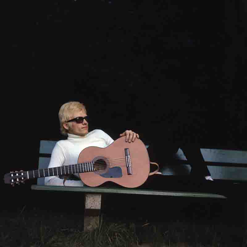 Der Sänger Heino entspannt liegend auf einer grünen Parkbank mit einer Akustikgitarre, weißem Rollkragenpullover und dunkler Sonnenbrille. Foto von Ingeborg Bock-Schroeder.