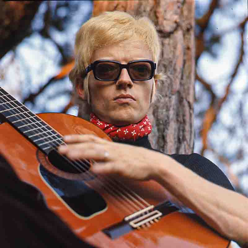 Nahaufnahme des jungen Sängers Heino im Freien vor einem Baumstamm. Er trägt sein charakteristisches hellblondes Haar, eine markante dunkle Brille und ein rotes Halstuch mit weißem Muster zu einem schwarzen Oberteil. Im Vordergrund ist eine Akustikgitarre angeschnitten, auf deren Saiten seine Hand ruht, fotografiert von Ingeborg Bock-Schroeder.