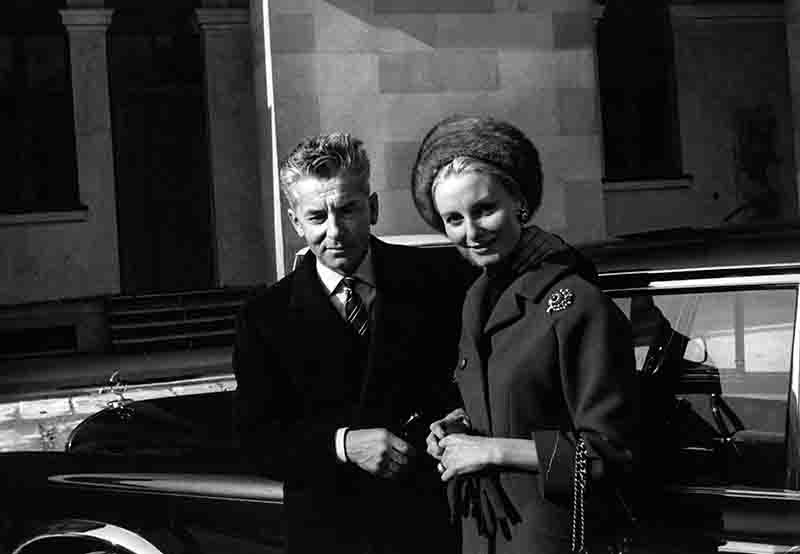 Schwarz-Weiß-Aufnahme des Dirigenten Herbert von Karajan und seiner Frau Eliette, elegant gekleidet vor einer klassischen Mercedes-Benz Limousine stehend.