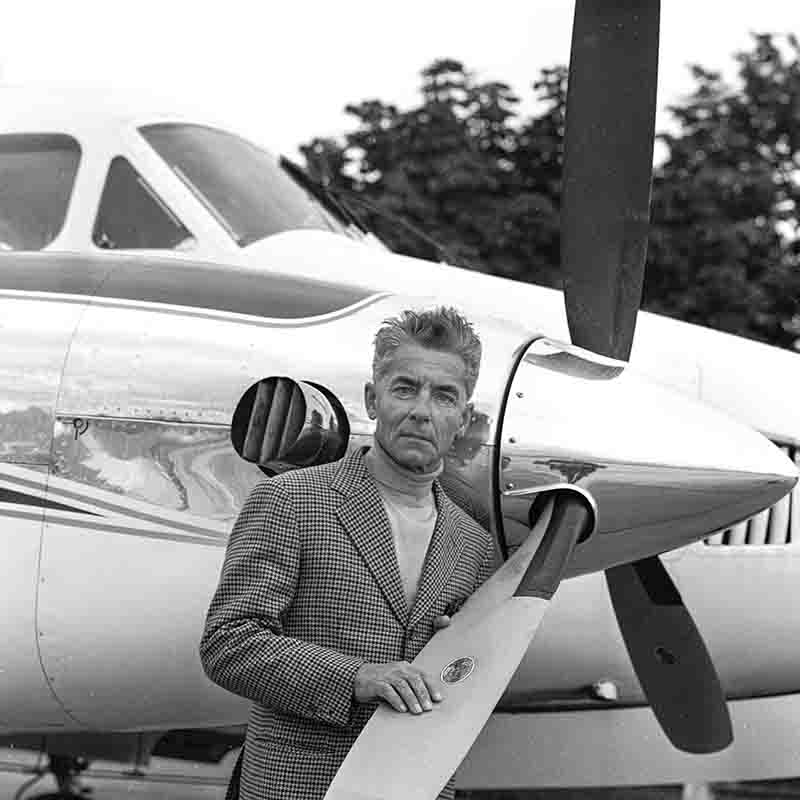Schwarz-Weiß-Porträt des Dirigenten Herbert von Karajan vor einem zweimotorigen Propellerflugzeug, eine Hand auf den Propeller gelegt.