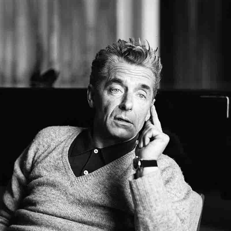 Schwarz-Weiß-Nahaufnahme von Herbert von Karajan im grauen Strickpullover, die Finger leicht an die Wange gelegt, mit direktem Blick.