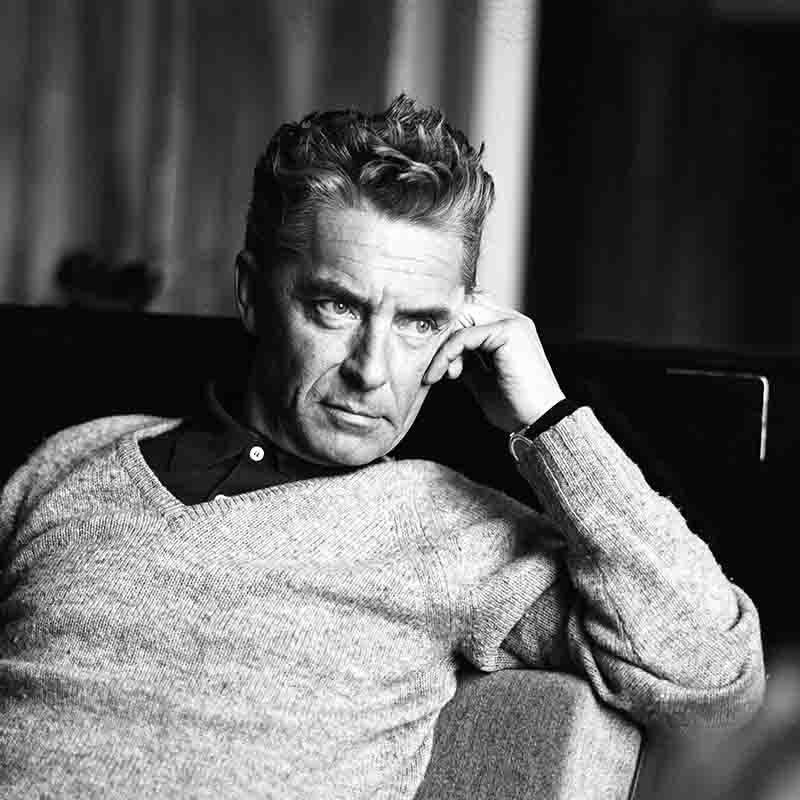 Schwarz-Weiß-Porträt des Dirigenten Herbert von Karajan, der den Kopf auf seine Hand stützt und einen strukturierten Strickpullover mit V-Ausschnitt über einem dunklen Hemd trägt.