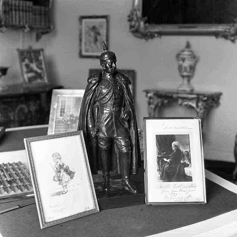 Schwarz-Weiß-Fotografie eines Ensembles historischer Erinnerungsstücke: Im Zentrum steht eine Bronzestatue von Kaiser Wilhelm II. mit Pickelhaube und Umhang, flankiert von zwei gerahmten Porträts, links eine Kinderzeichnung, rechts eine Fotografie einer älteren Dame mit handschriftlicher Widmung.