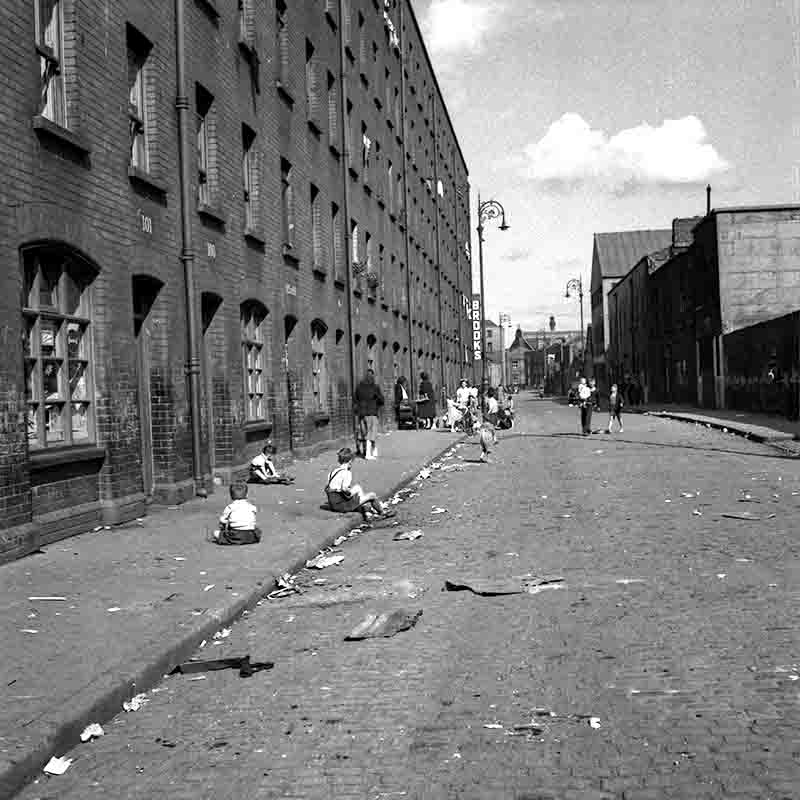 Schwarz-Weiß-Fotografie von Peter Bock-Schroeder aus Dublin: Eine kopfsteingepflasterte Straße flankiert von einer langen Backsteinhäuserzeile. Auf dem Gehweg sitzen und spielen mehrere Kinder, während im Hintergrund Passanten zu sehen sind und leichtes Müllpapier auf dem Straßenbelag liegt.