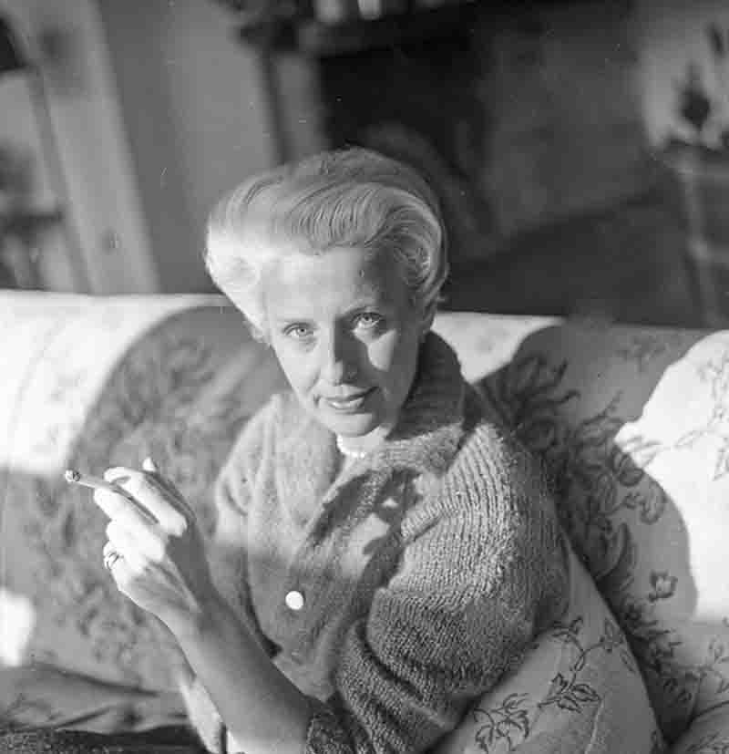 Schwarz-weiß Porträt der Modedesignerin Maria Bogner, fotografiert von Ingeborg Bock-Schroeder. Sie sitzt auf einem gemusterten Sofa, trägt eine helle Strickjacke mit Perlenkette und hält eine brennende Zigarette in der Hand. Das Licht fällt seitlich auf ihr Gesicht und betont ihre elegante Erscheinung.