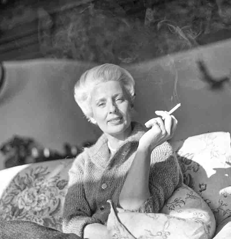 Schwarz-weiß Porträtaufnahme von Maria Bogner, fotografiert von Ingeborg Bock-Schröder. Die Modedesignerin sitzt auf einem floral gemusterten Sofa und hält eine brennende Zigarette empor, deren Rauch im Gegenlicht deutlich sichtbar ist. Sie trägt eine helle Strickjacke und blickt entspannt in die Kamera.