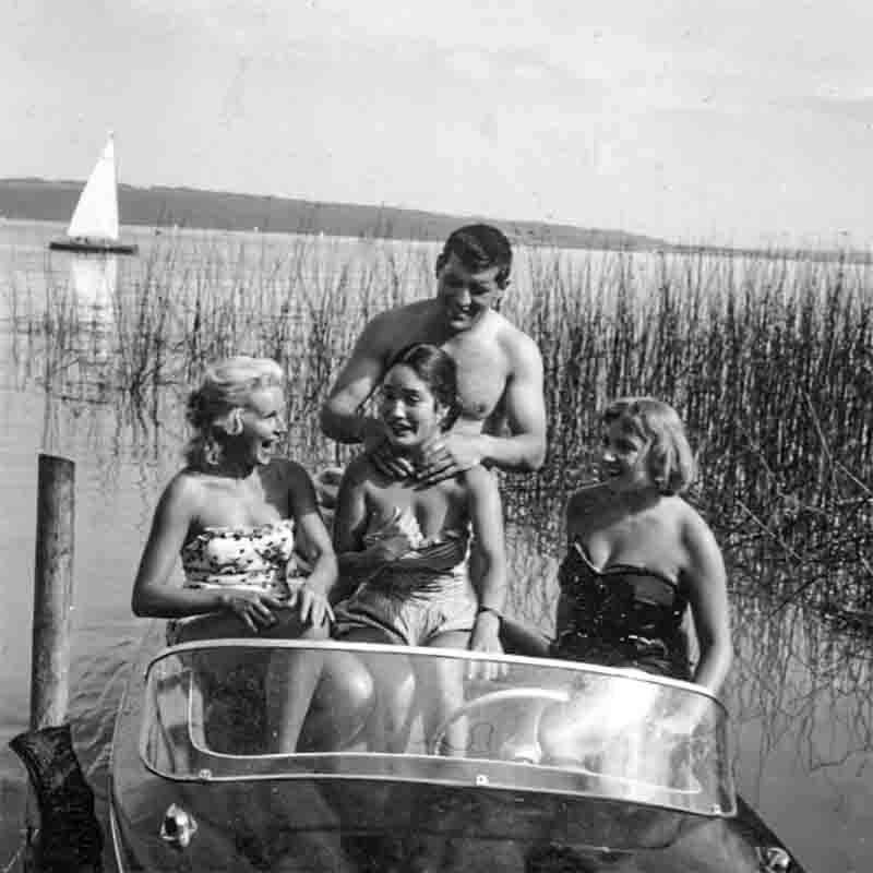 Historische Schwarz-Weiß-Aufnahme von Mario Adorf mit Ellen Schwiers und Freundinnen in Badekleidung auf einem Motorboot. Die Gruppe lacht und genießt einen Sommertag auf einem See, im Hintergrund ist ein Segelboot und Schilf zu sehen. Foto von Ingeborg Bock-Schroeder.