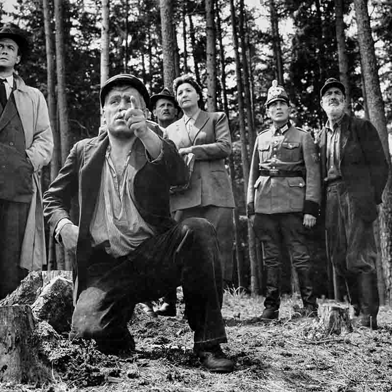 Schwarz-Weiß-Szenenfoto von Mario Adorf am Set des Films „Nachts, wenn der Teufel kam“, fotografiert von Ingeborg Bock-Schroeder. Adorf kniet im Wald und zeigt mit ausgestrecktem Finger nach oben, umgeben von weiteren Darstellern und Statisten in historischen Kostümen.