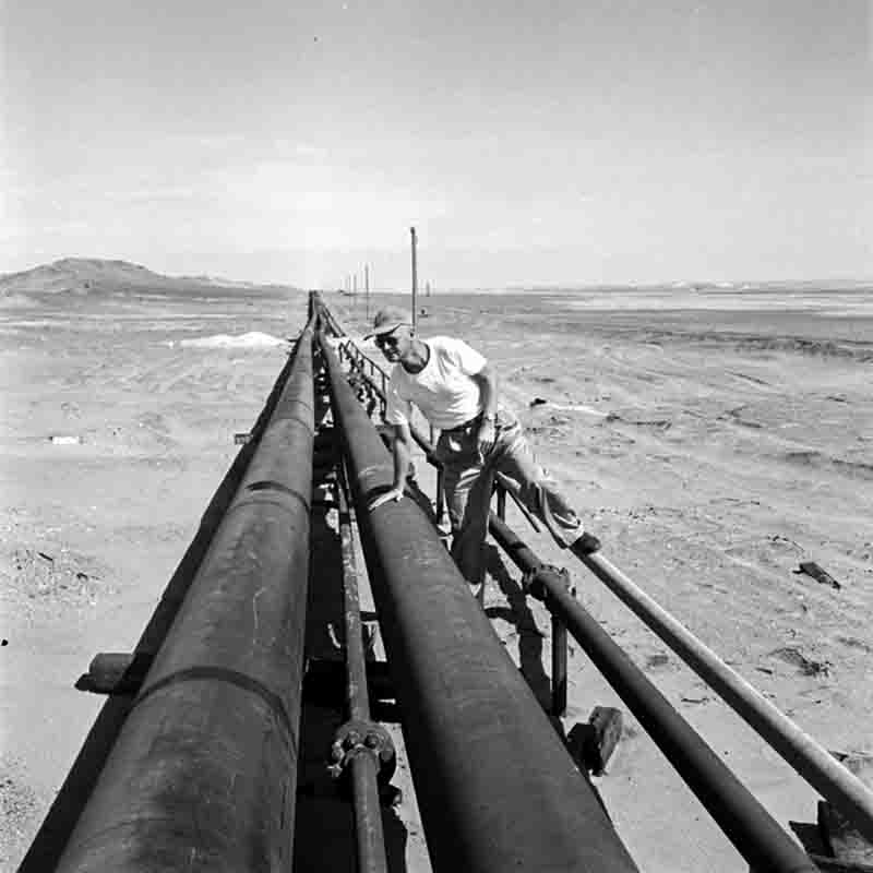 Schwarz-Weiß-Fotografie von Peter Bock-Schroeder aus der Talara-Wüste in Peru: Ein Mann mit Kappe und Sonnenbrille lehnt an einer langen Pipeline, die sich schnurgerade durch die weite, karge Wüstenlandschaft bis zum Horizont erstreckt.