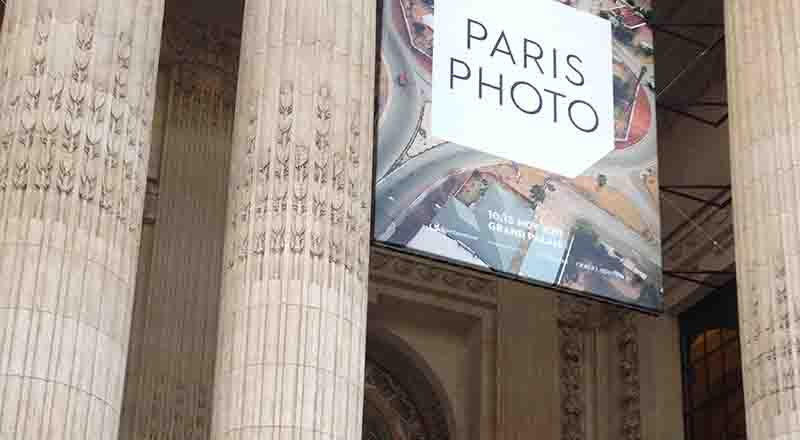 Ein großes Werbebanner für die Kunstmesse „PARIS PHOTO“ am Grand Palais, aufgehängt zwischen klassischen, verzierten Steinsäulen. Das Banner zeigt ein Luftbild einer Landschaft sowie den Schriftzug „PARIS PHOTO“ und Veranstaltungsdetails.