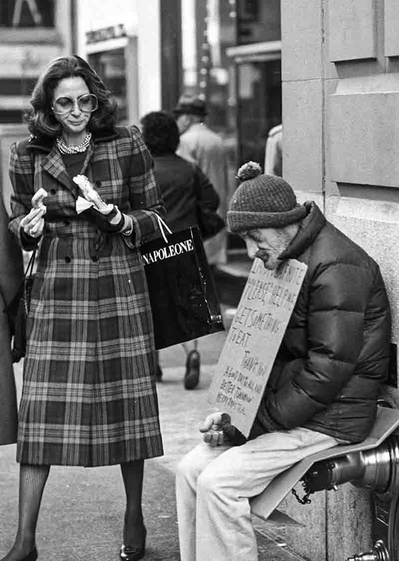 Schwarz-Weiß-Fotografie von Peter Bock-Schroeder aus der New York 1982 Serie: Eine elegant gekleidete Frau im karierten Mantel mit Sonnenbrille und Napoleone-Einkaufstasche geht an einem obdachlosen Mann mit Pappschild vorbei, der auf der Straße sitzt.