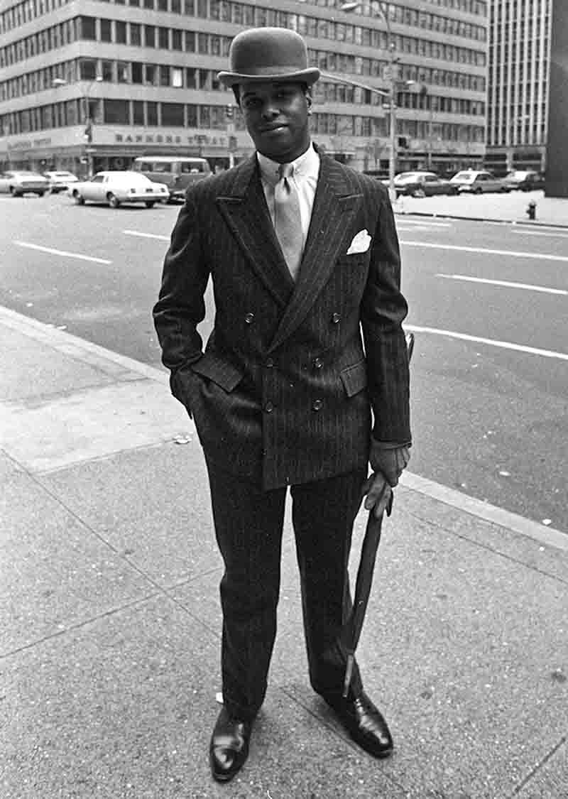 Schwarz-Weiß-Fotografie von Peter Bock-Schroeder aus der New York 1982 Serie: Porträt eines elegant gekleideten afroamerikanischen Mannes auf einem Gehweg in Manhattan mit Nadelstreifenanzug, Melone (Bowler Hat), Krawatte, Einstecktuch und Regenschirm in der Hand vor einer Kulisse aus Straße und Bankgebäuden.