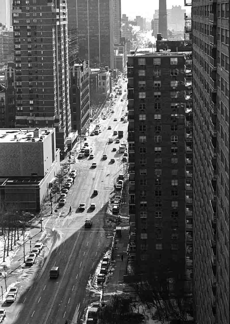 Schwarz-Weiß-Fotografie von Peter Bock-Schroeder aus der New York 1982 Serie: Vogelperspektive auf eine belebte Avenue in Manhattan mit fahrenden Autos, Schattenwurf hoher Wolkenkratzer und leicht verschneiten Bürgersteigen.