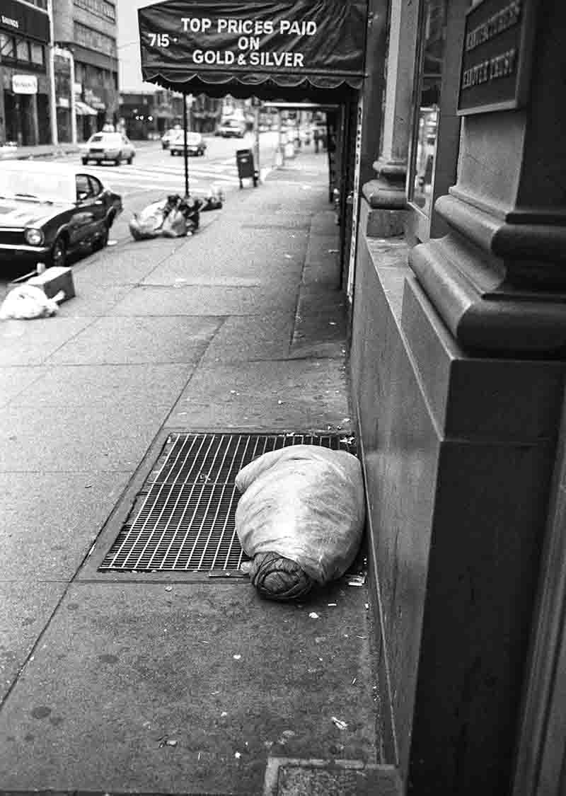 Schwarz-Weiß-Fotografie von Peter Bock-Schroeder aus der New York 1982 Serie: Eine obdachlose Person schläft in Plastik eingewickelt auf einem Lüftungsgitter auf dem Gehweg vor einem Geschäft mit der Aufschrift „TOP PRICES PAID ON GOLD & SILVER“.