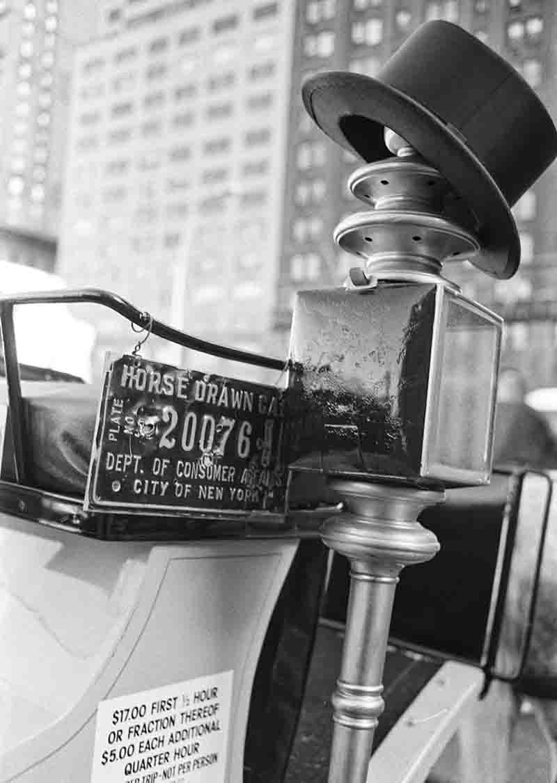 Schwarz-Weiß-Fotografie von Peter Bock-Schroeder aus der New York 1982 Serie: Nahaufnahme einer traditionellen Pferdekutsche mit einem Zylinderhut auf einer Laterne, einem offiziellen Lizenzschild und Preisaushang vor verschwommenen Hochhäusern.