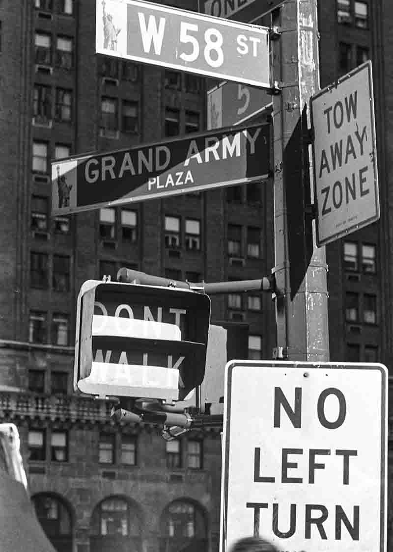 Schwarz-Weiß-Fotografie von Peter Bock-Schroeder aus der New York 1982 Serie: Nahaufnahme von New Yorker Straßenschildern für West 58th Street und Grand Army Plaza sowie Schildern für „DONT WALK“, „NO LEFT TURN“ und „TOW AWAY ZONE“ vor einer Gebäudefassade.