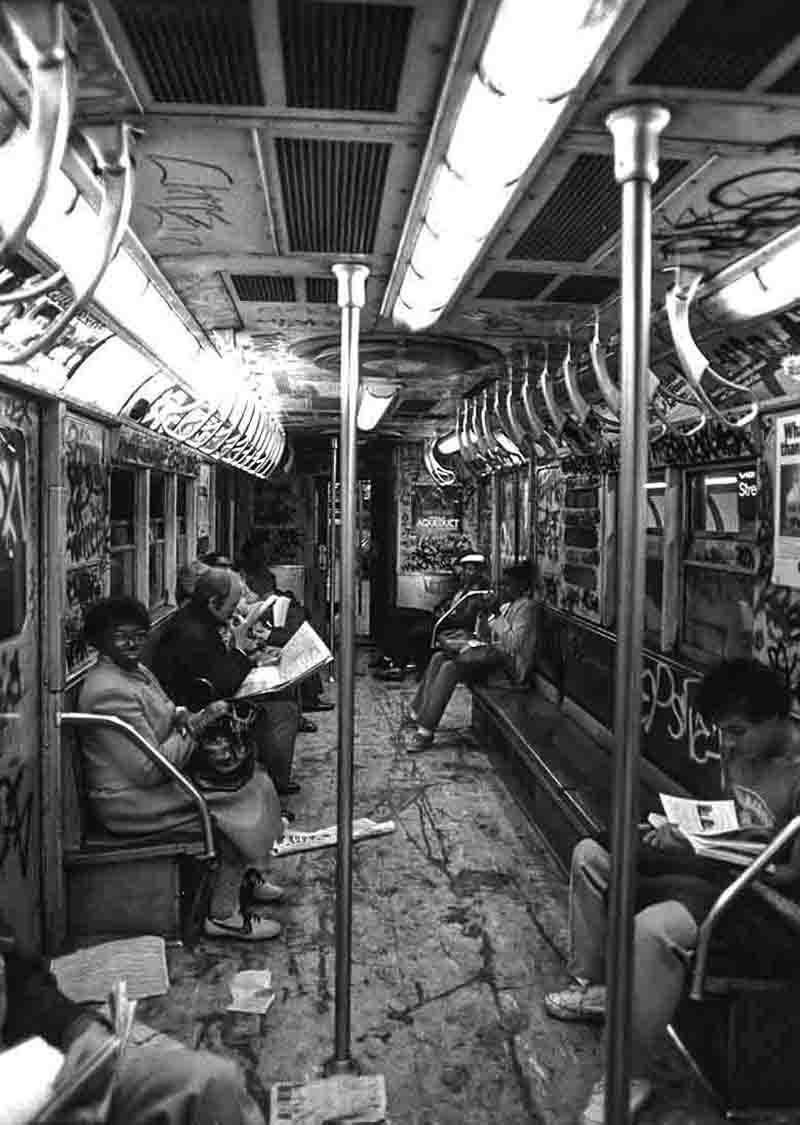 Schwarz-Weiß-Fotografie von Peter Bock-Schroeder aus der New York 1982 Serie: Das Innenleben eines von Graffitis übersäten New Yorker U-Bahn-Wagons, in dem Fahrgäste sitzen und Zeitung lesen.