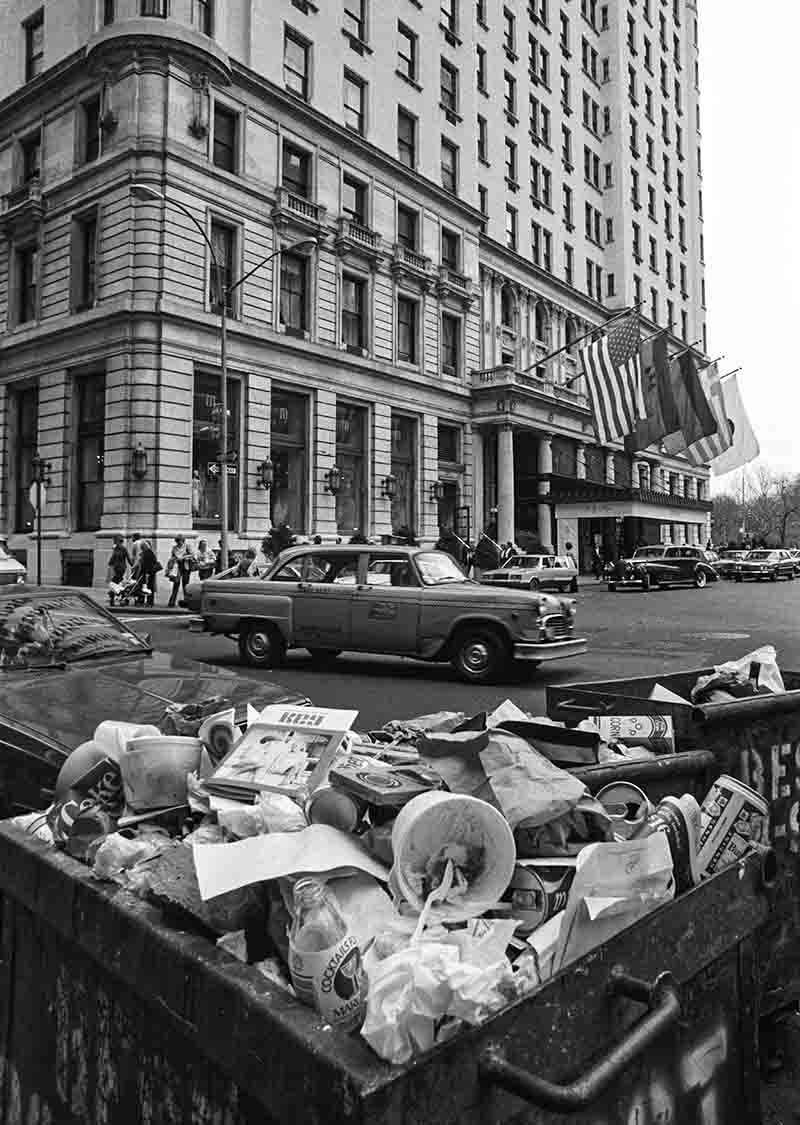 Schwarz-Weiß-Fotografie von Peter Bock-Schroeder aus dem Jahr 1982 in New York City: Im Vordergrund steht ein überquellender Müllcontainer voller Abfall, Becher und Flaschen, während im Hintergrund das elegante Hotel Waldorf Astoria mit US-Flaggen und ein vorbeifahrendes Taxi zu sehen sind.