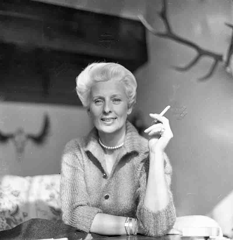 Schwarz-weiß Porträt von Maria Bogner, fotografiert von Ingeborg Bock-Schroeder. Die Modedesignerin blickt lächelnd in die Kamera, trägt eine helle Strickjacke, eine Perlenkette und hält eine brennende Zigarette in der Hand. Im Hintergrund sind Geweihe an der Wand zu sehen.