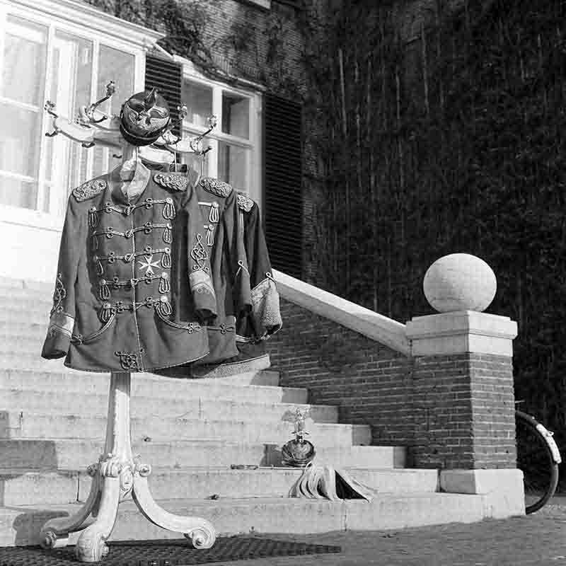 Historische Schwarz-Weiß-Fotografie von Peter Bock-Schroeder (1960): Eine preußische Husarenuniform mit markanter Schnürung und Epauletten sowie eine Pickelhaube hängen an einem Kleiderständer im Außenbereich von Haus Doorn. Die Uniformen sind vor der Freitreppe des Schlosses platziert.