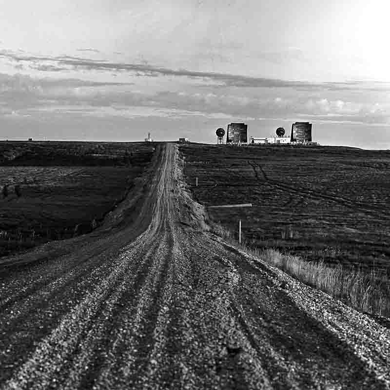 Schwarz-Weiß-Fotografie von Peter Bock-Schroeder aus Alaska: Eine unbefestigte Kiesstraße führt geradlinig durch eine weite, flache Tundralandschaft auf eine abgelegene Radarstation mit großen Antennen und Parabolspiegeln am Horizont zu.