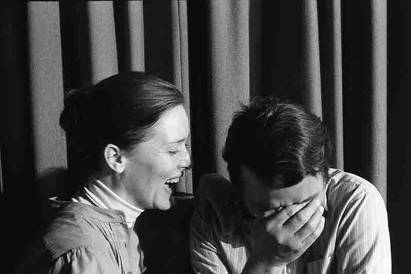 Schwarz-weißes Selbstporträt von Peter und Ingeborg Bock-Schröder vor einem dunklen Vorhang. Ingeborg lacht herzlich im Profil, während Peter rechts daneben sitzt und sich amüsiert das Gesicht in die Hand stützt. Die Szene wirkt privat und ist durch starke Licht-Schatten-Kontraste geprägt.