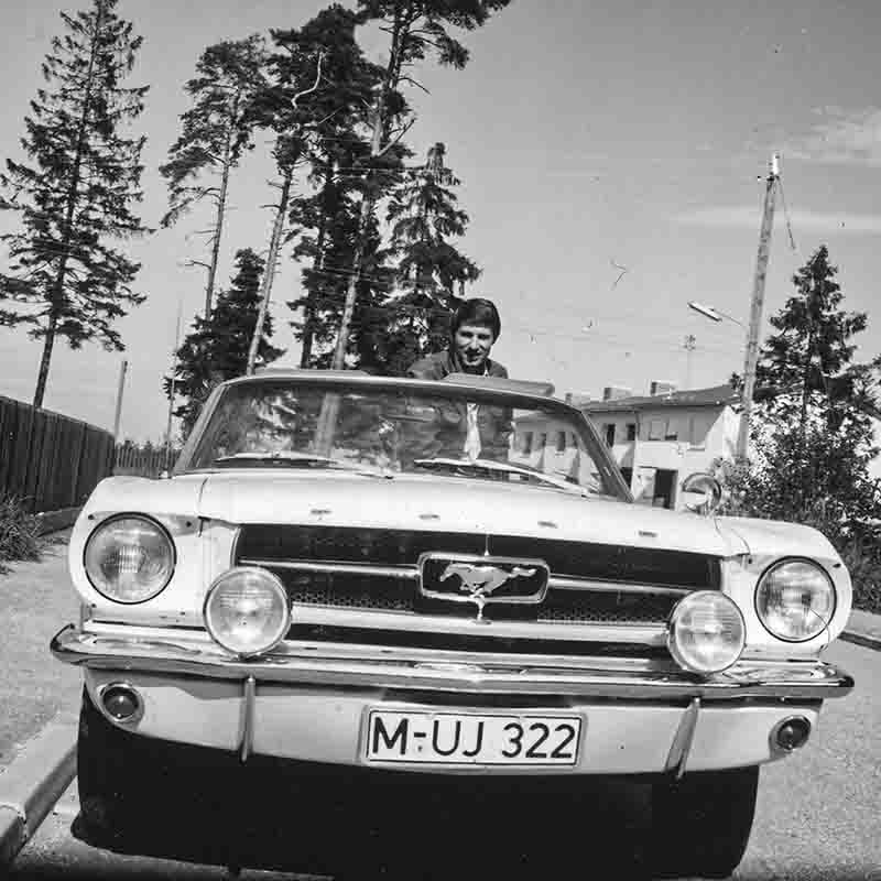 Schwarz-Weiß-Fotografie von Udo Jürgens, der am Steuer eines klassischen Ford Mustang Cabriolets sitzt. Das Auto hat das Münchner Kennzeichen M-UJ 322. Die Aufnahme von Ingeborg Bock-Schroeder zeigt den Künstler lächelnd vor einer Kulisse aus hohen Nadelbäumen und Gebäuden.