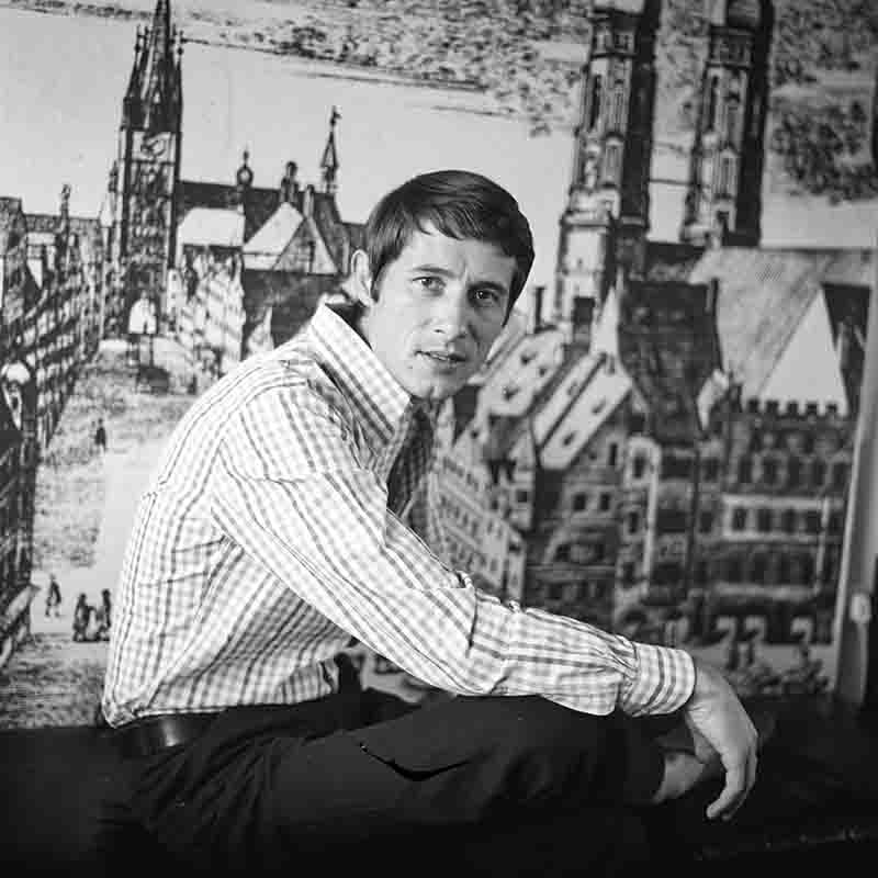 Schwarz-Weiß-Porträt eines jungen Udo Jürgens, fotografiert von Ingeborg Bock-Schroeder. Er sitzt im Profil vor einer großflächigen Wandgrafik der Münchner Frauenkirche, trägt ein helles kariertes Hemd und blickt direkt in die Kamera.